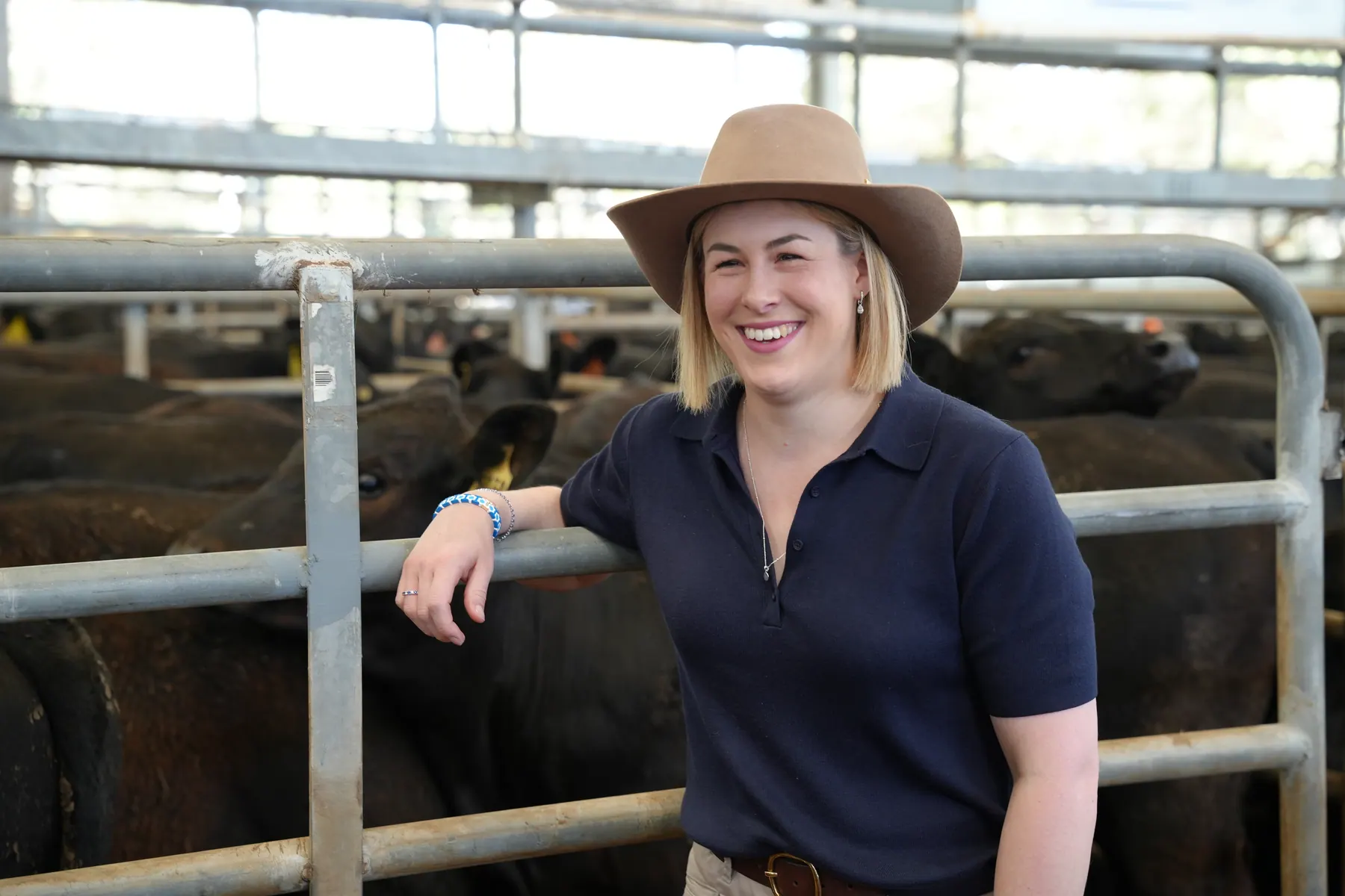 Jess Wilson smiling at a regional saleyard, leaning on a cattle rail in a wide-brimmed hat